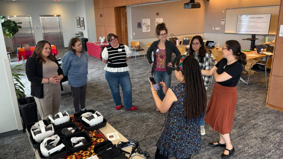 Group of participants standing in a classroom around a table with virtual reality headsets. One person explains instructions while holding a phone, as others listen. A screen in the background displays VR and AR safety guidelines.